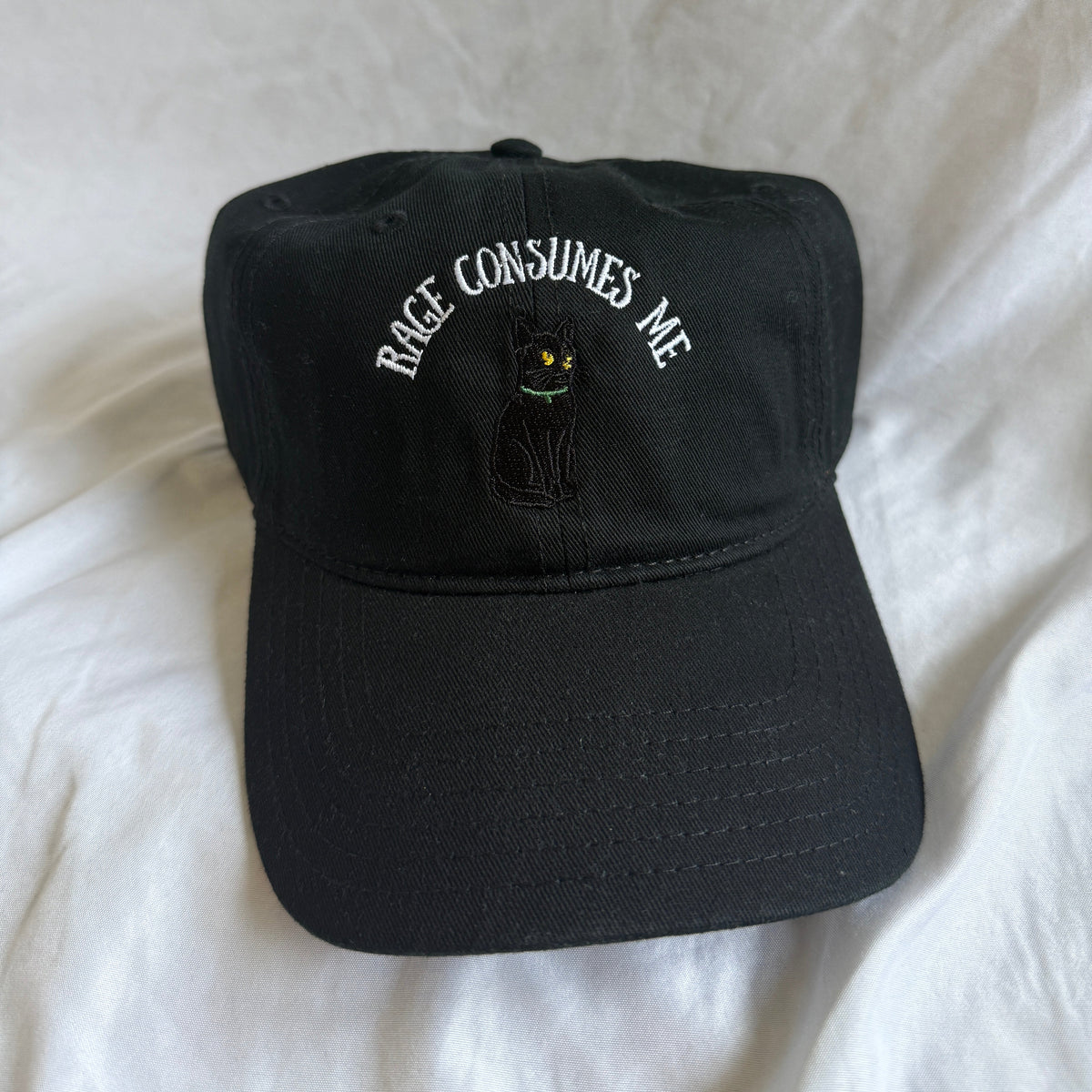 A black baseball cap with white embroidered text reading 'RACE CONSUMES ME' and a small black cat graphic, resting on a white fabric background.