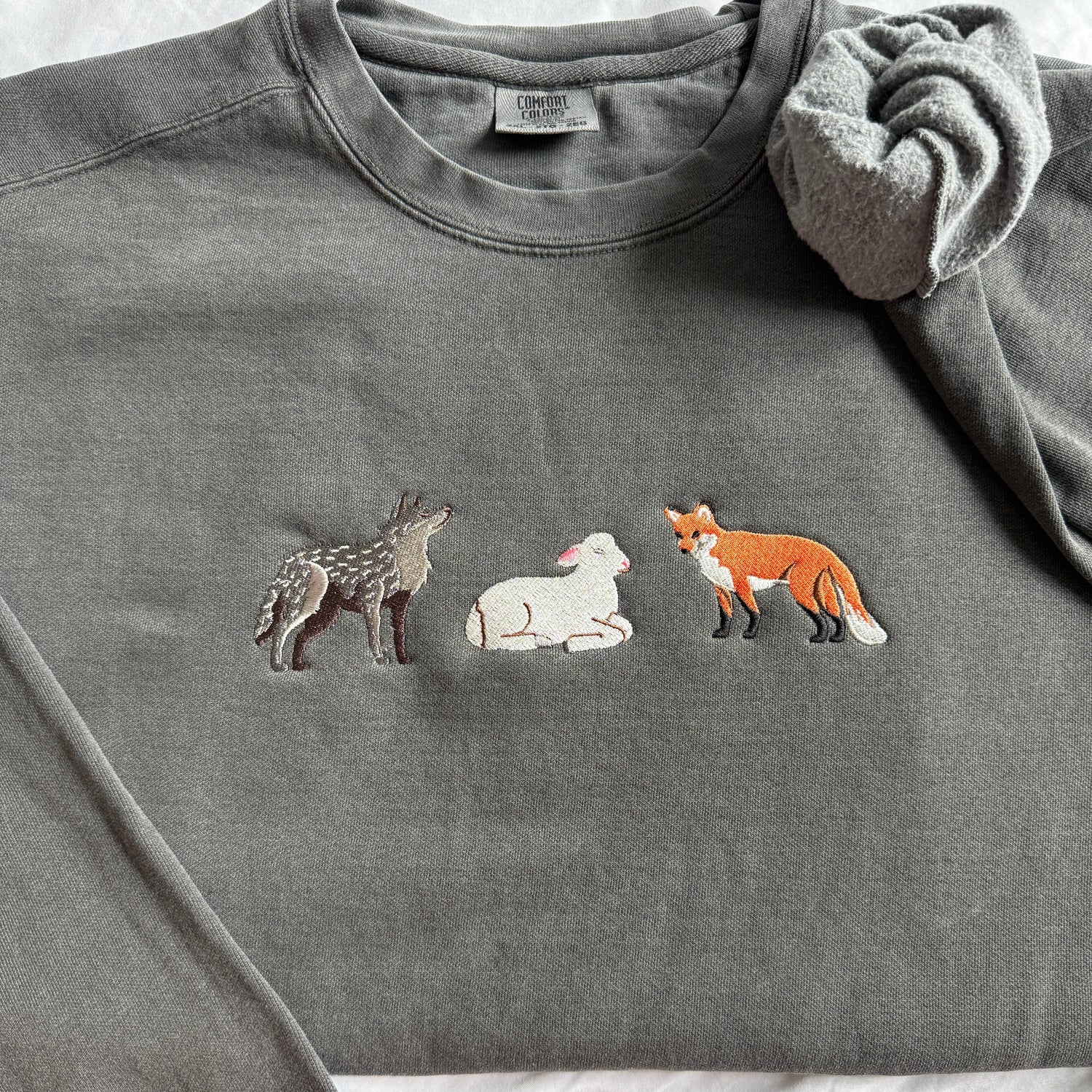 A dark gray crewneck sweatshirt featuring embroidered animal characters—a wolf, a white lamb, and a fox—on the chest, styled flat on a white fabric background.