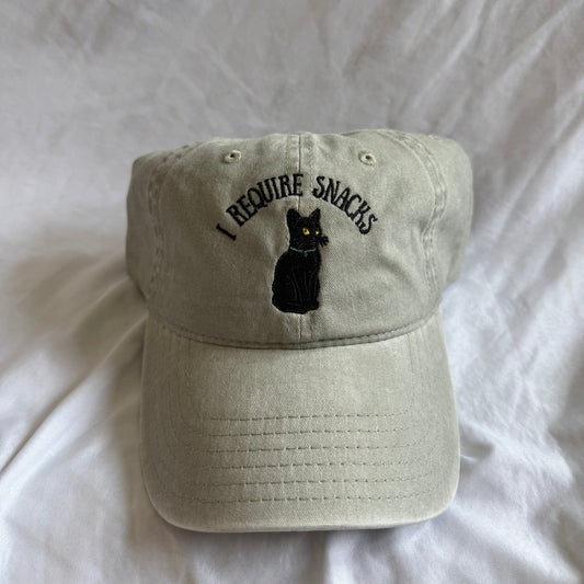 A light beige baseball cap with 'I REQUIRE SNACKS' embroidered in black above a black cat graphic with yellow eyes, resting on a white fabric background.