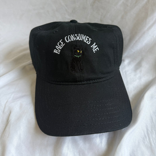 A black baseball cap with white embroidered text reading 'RACE CONSUMES ME' and a small black cat graphic, resting on a white fabric background.