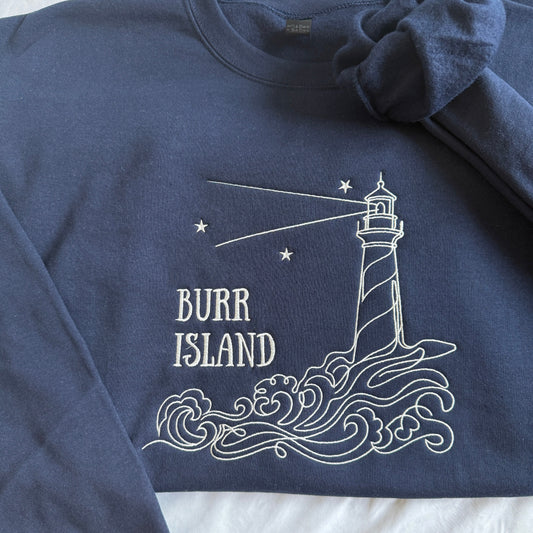 A navy blue crewneck sweatshirt featuring a white line art graphic of a lighthouse on waves with the text 'BURR ISLAND' and small stars. The sweatshirt is laid flat on a white fabric background with a matching navy knit scrunchie tied to the collar.