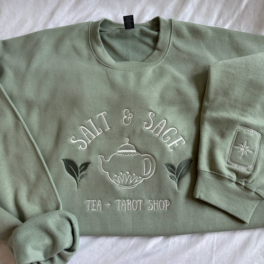 A sage green crewneck sweatshirt with a white embroidered graphic reading 'SALT & SAGE TEA + TAROT SHOP' and a teapot illustration, laid flat on a white fabric background.