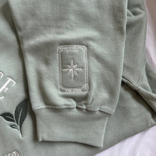 A close-up of a sage green sweatshirt sleeve featuring a rectangular embroidered patch with a compass rose design and the text 'THE STAR' on the cuff.