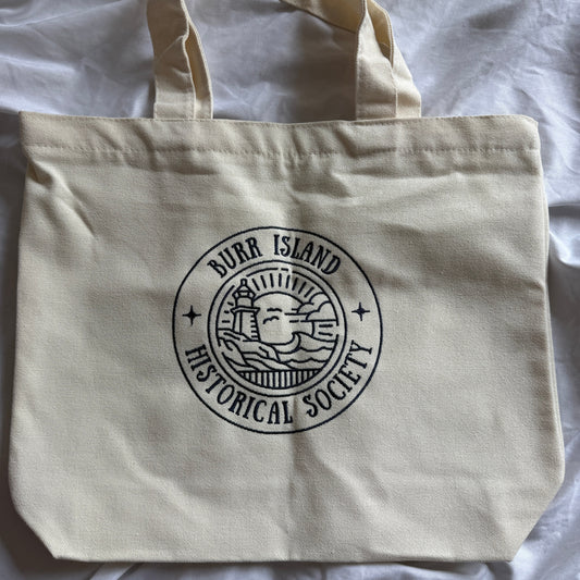 A natural canvas tote bag with a dark gray printed circular seal reading 'Burr Island Historical Society' laid flat on a wrinkled white sheet.
